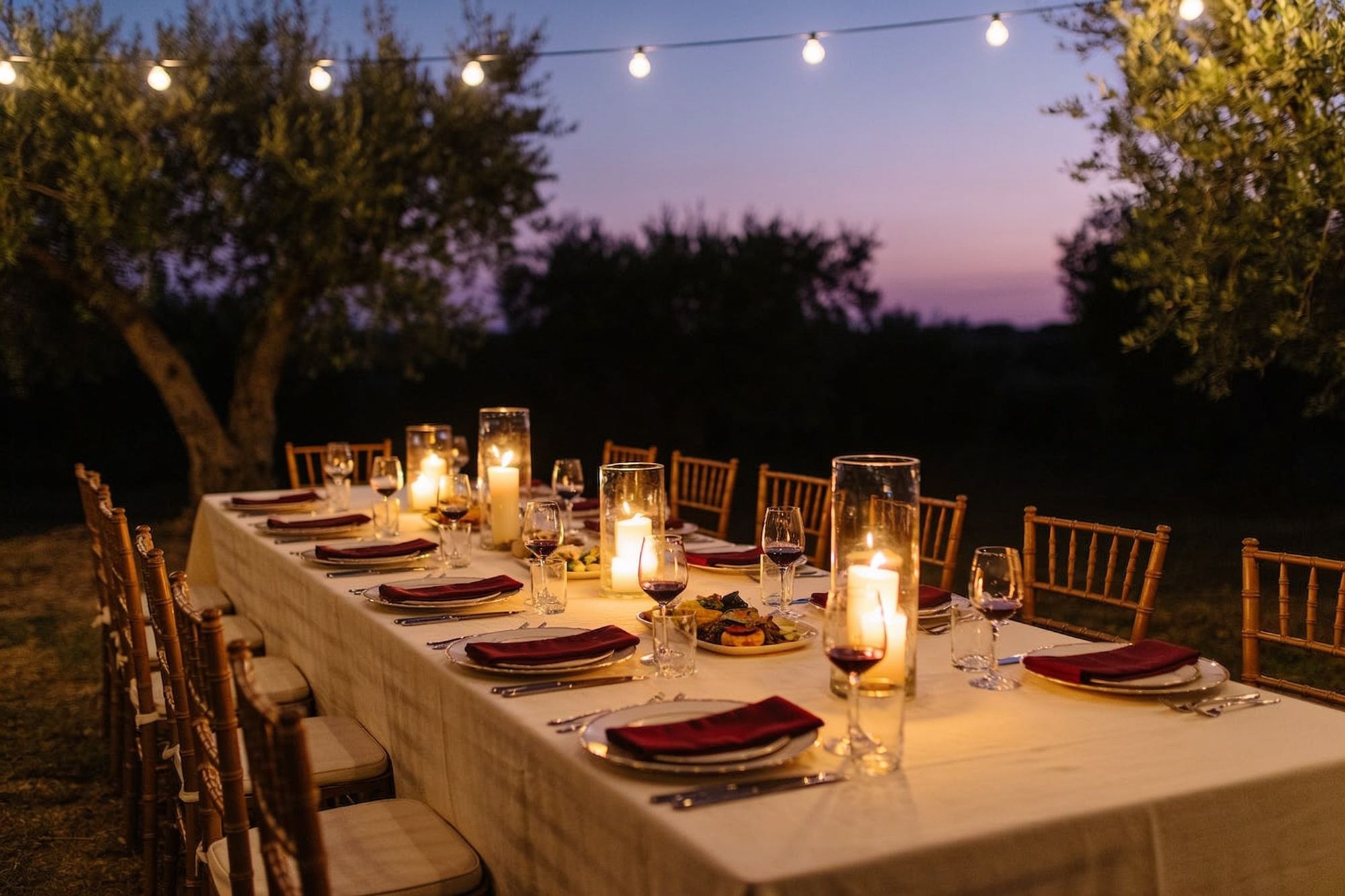 A long dinner table under string lights and olive trees at dusk